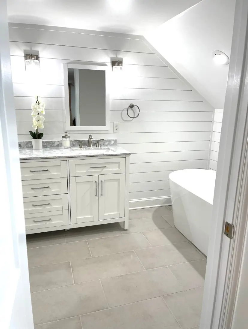 A modern white bathroom with a vanity, mirror, freestanding tub, and shiplap walls under soft lighting.