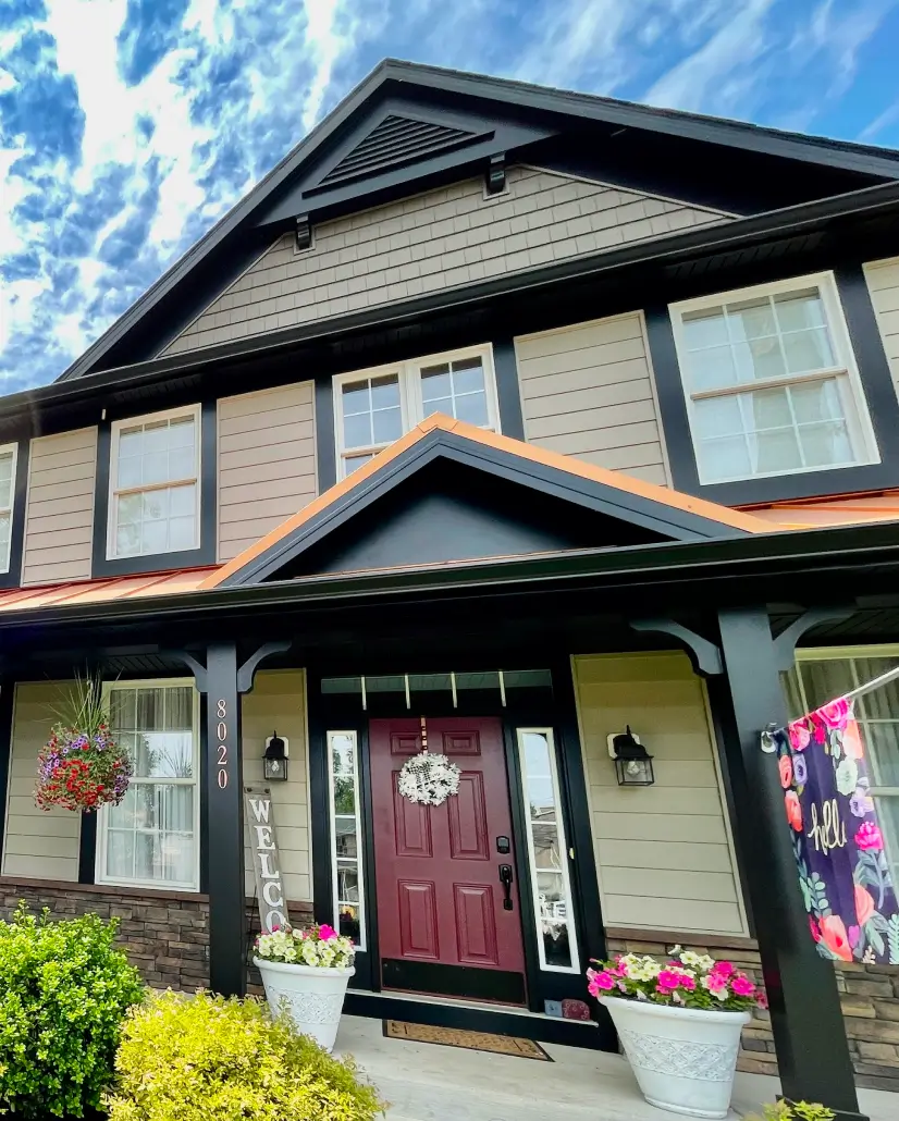Two-story house with a red door, hanging flower baskets, and a welcome sign on a sunny day.