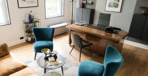 Modern office with teal chairs, wooden desk, wall art, and a small round table with a scale on it.