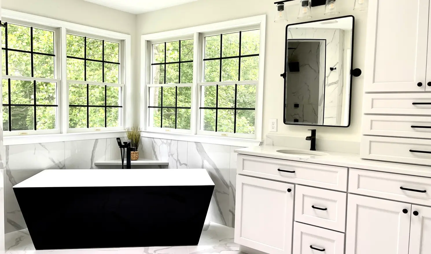 Modern bathroom with a black and white tub, white cabinets, large windows, and marble accents.