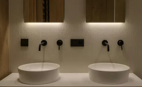 Modern bathroom with two round sinks, wall-mounted faucets, and illuminated mirrors above each sink.
