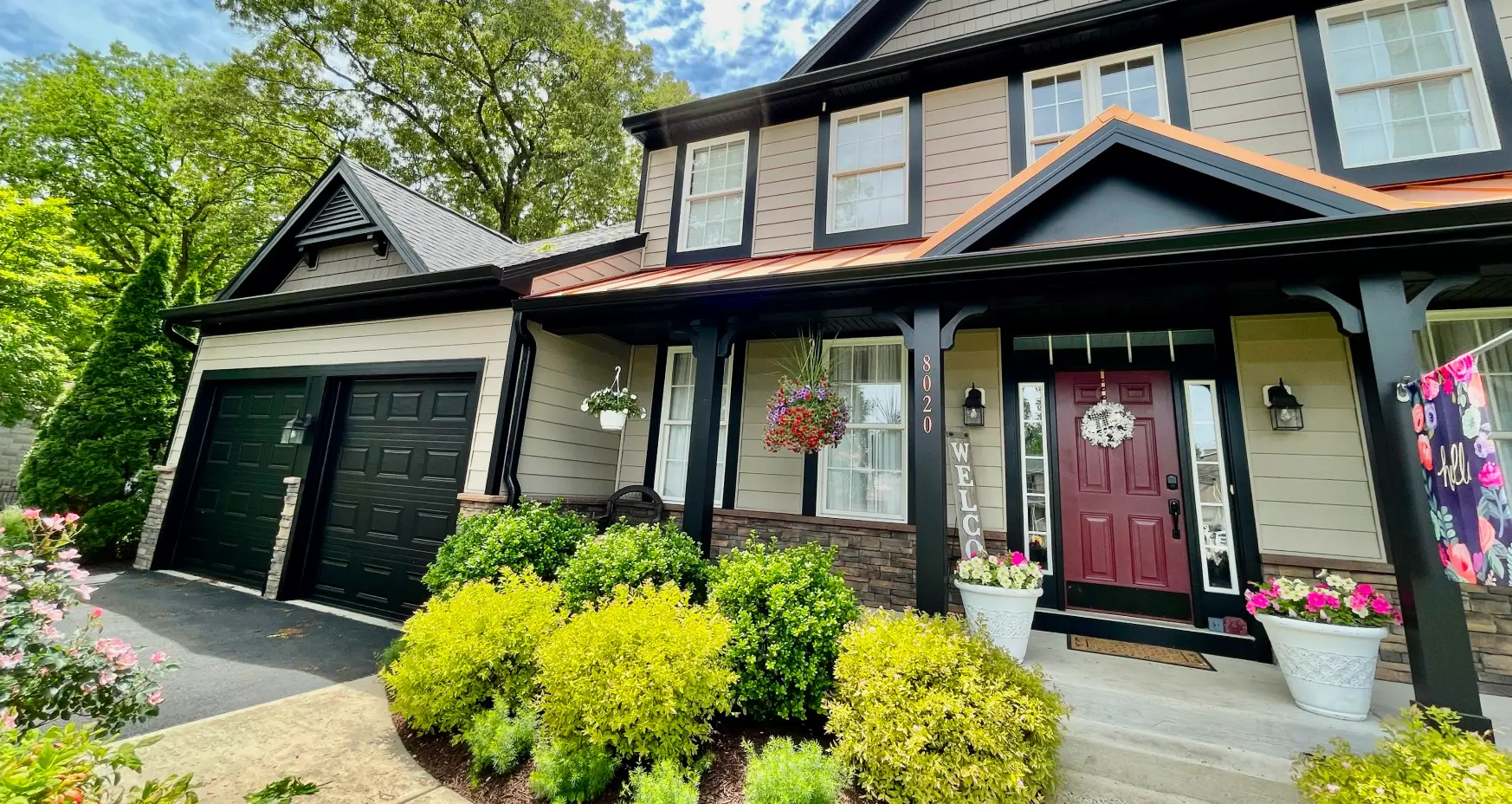 Charming two-story house with a red door, black garage, hanging plants, and green shrubs in front.