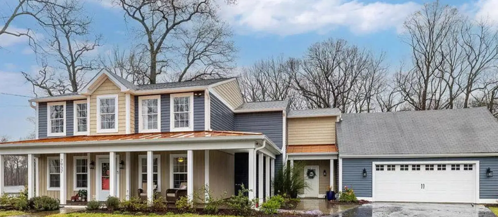 A two-story gray house with a porch and white garage, surrounded by leafless trees under a cloudy sky.