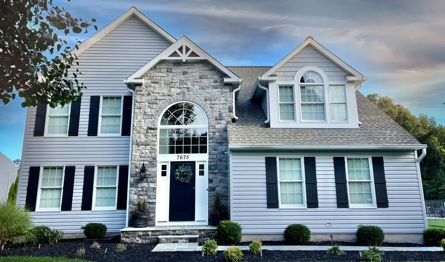 Two-story house with stone and siding exterior, arched window, dark shutters, and front garden under a cloudy sky.