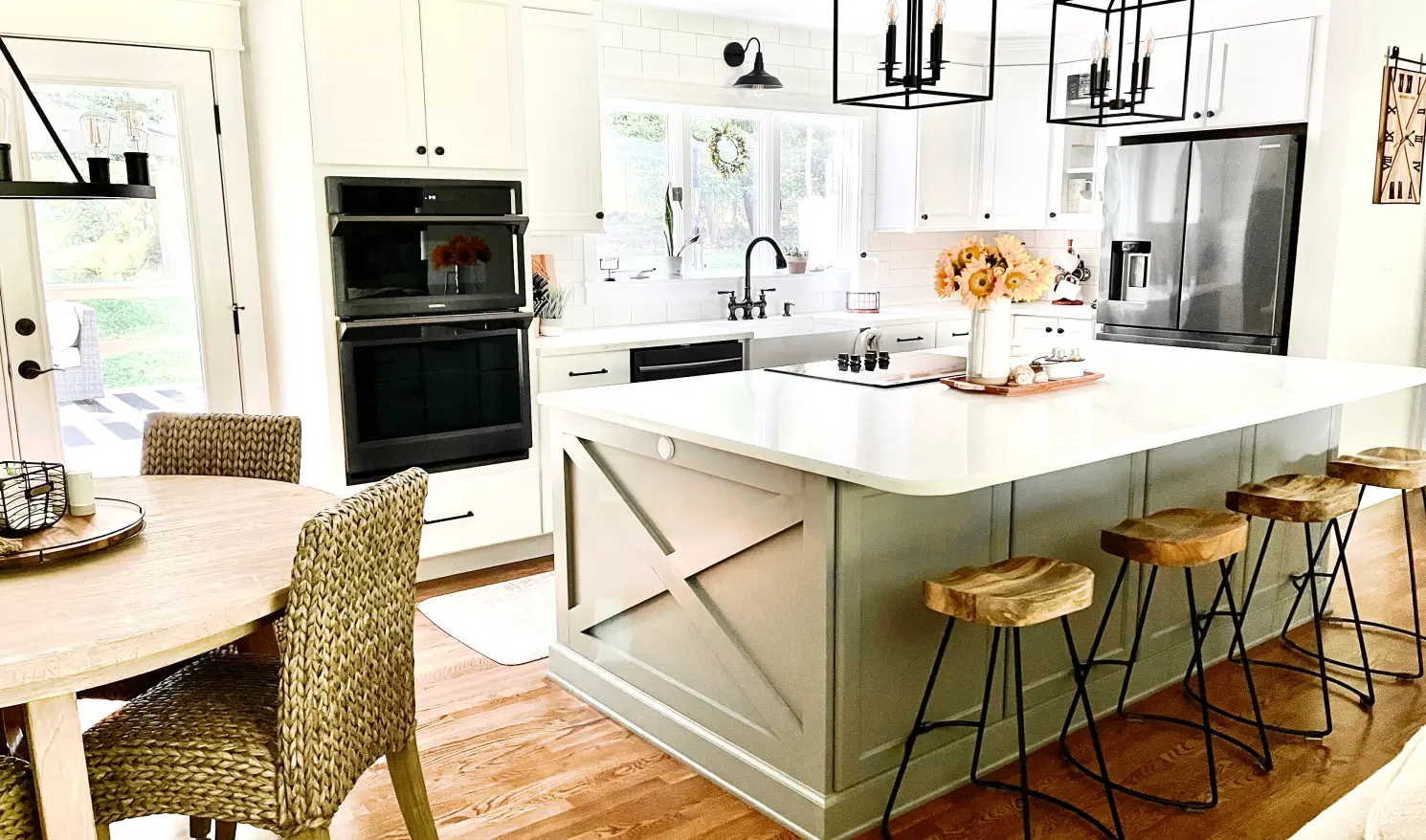 Bright kitchen with a large island, white cabinets, black appliances, and woven chairs around a wooden table.