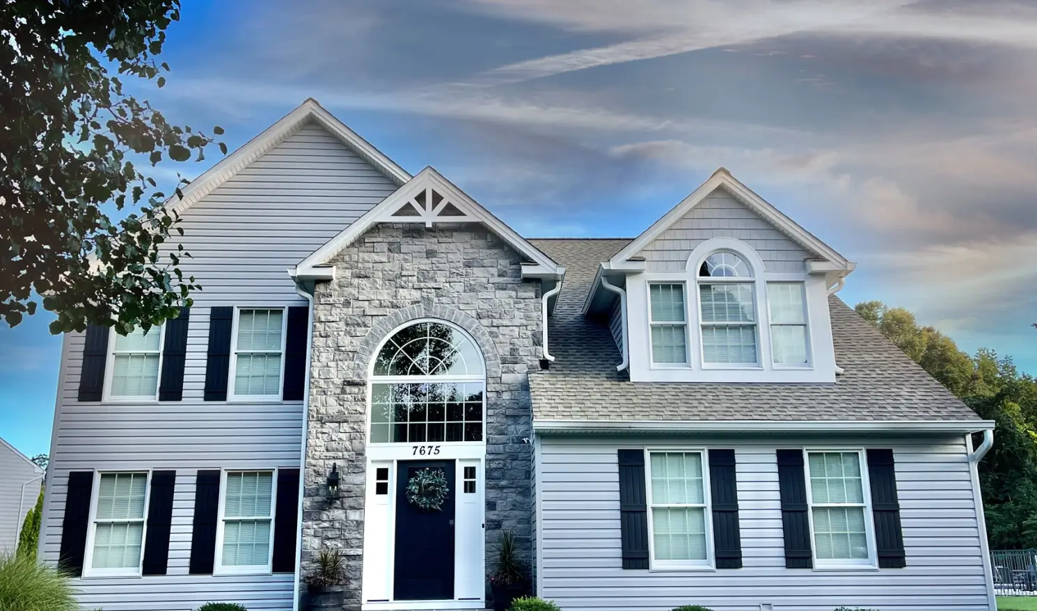 Two-story house with gray siding, stone facade, and black shutters under a blue sky with clouds.