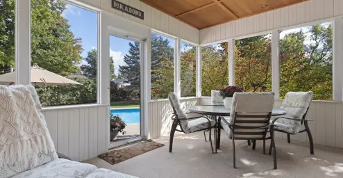 Bright sunroom with a table, chairs, and pool view through large windows. Trees and greenery outside.