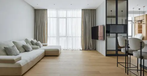 Minimalist living room with a cream sofa, large windows, and a TV on the wall. There are bar stools in the kitchen area.