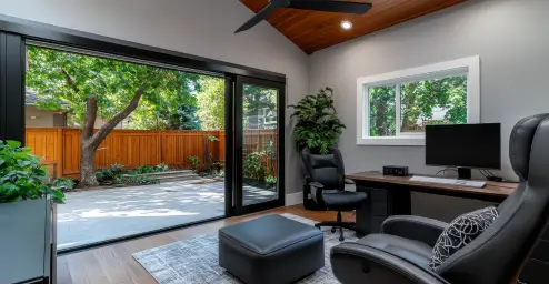 Modern home office with a desk, chair, and ottoman, opens to a patio with a wooden fence and trees in the background.