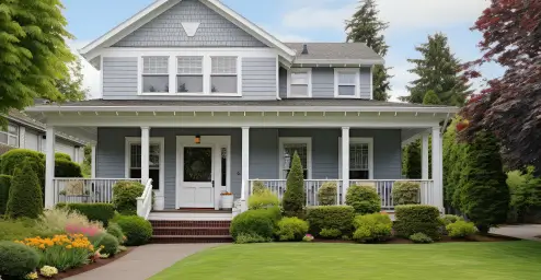 A two-story blue house with a porch, surrounded by lush greenery and colorful flowers.