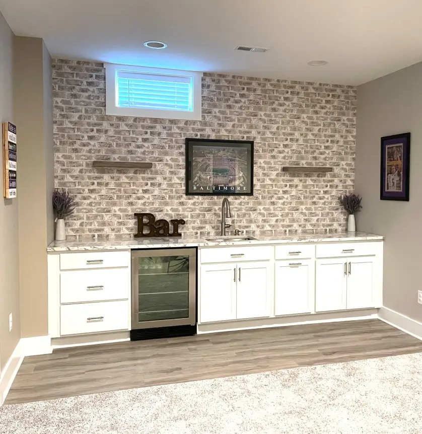 Basement bar with white cabinets, wine cooler, and faux brick backsplash, featuring bar sign and wall decor.