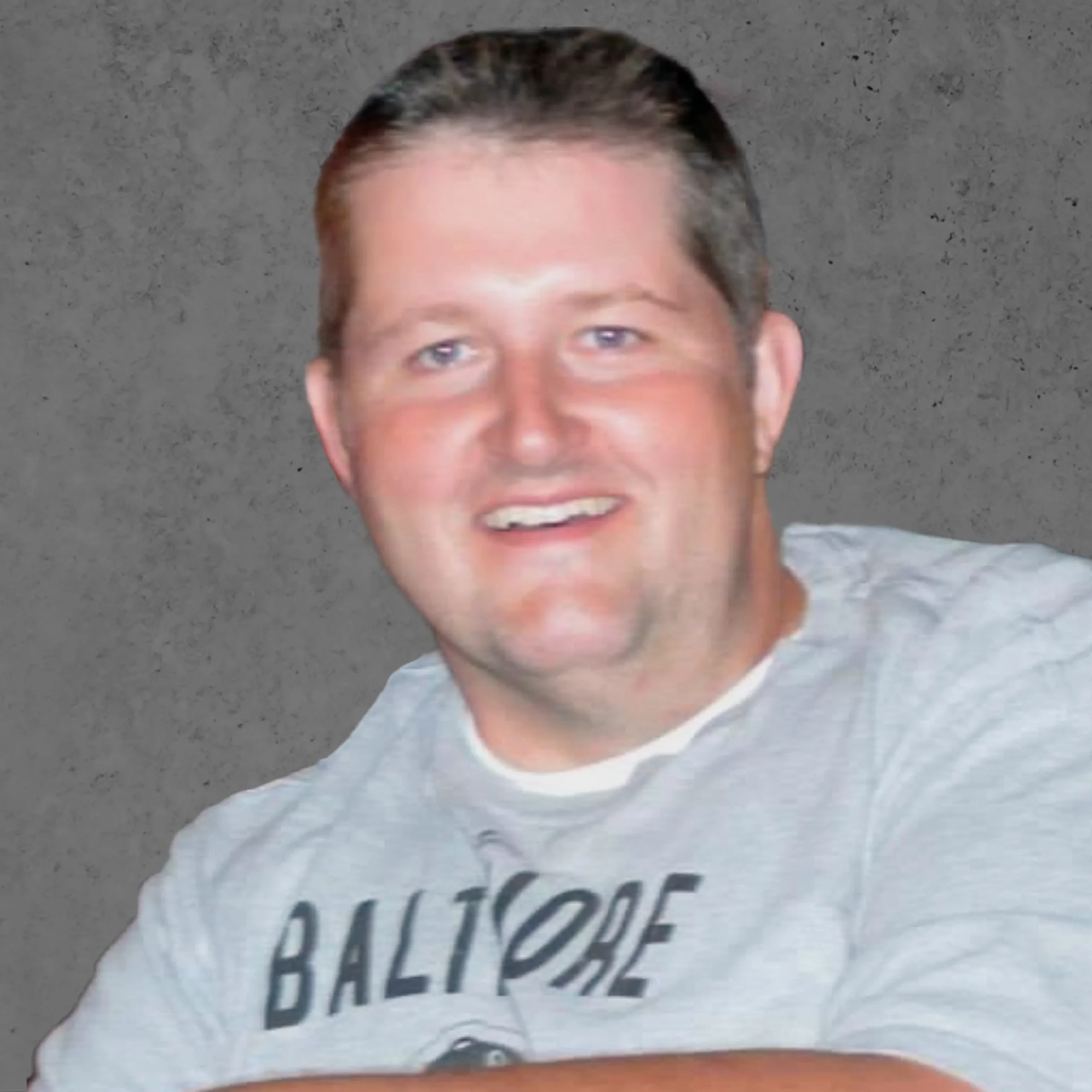 Smiling person wearing a gray "Baltimore" shirt, against a plain gray background.
