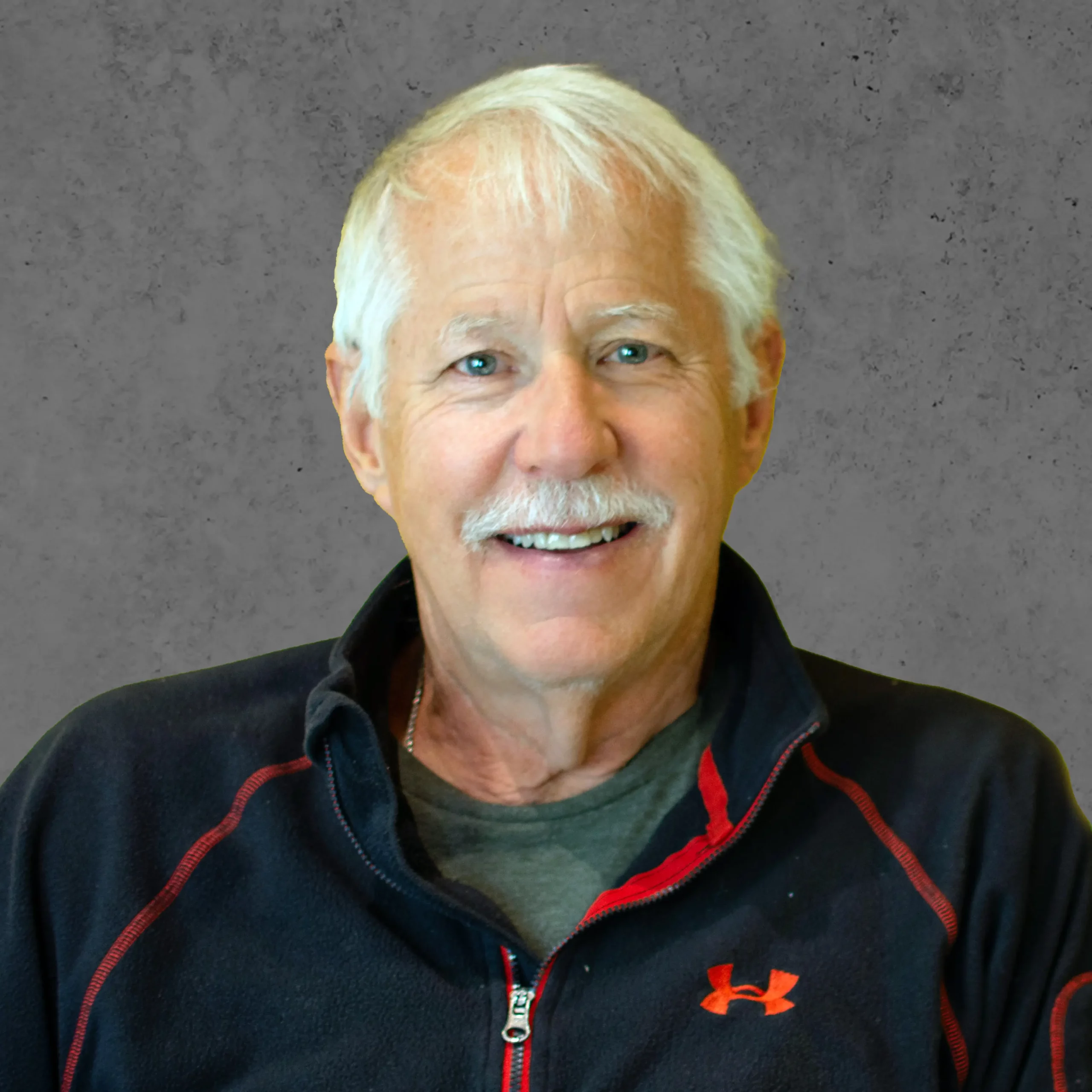 Older man with white hair and a mustache, wearing a black zip-up jacket with a red accent, smiling at the camera.