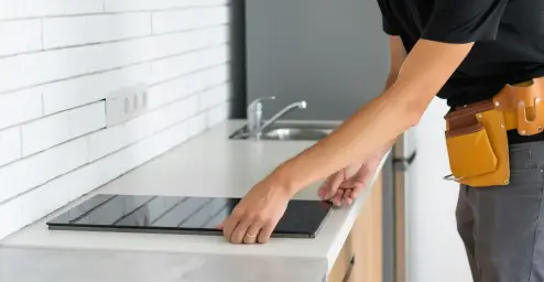 Person installing a modern, flat induction cooktop in a kitchen with a white countertop and brick-patterned backsplash.