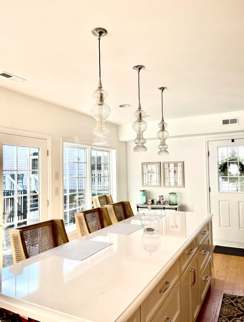Bright kitchen with a long white countertop, wicker chairs, pendant lights, and large windows.