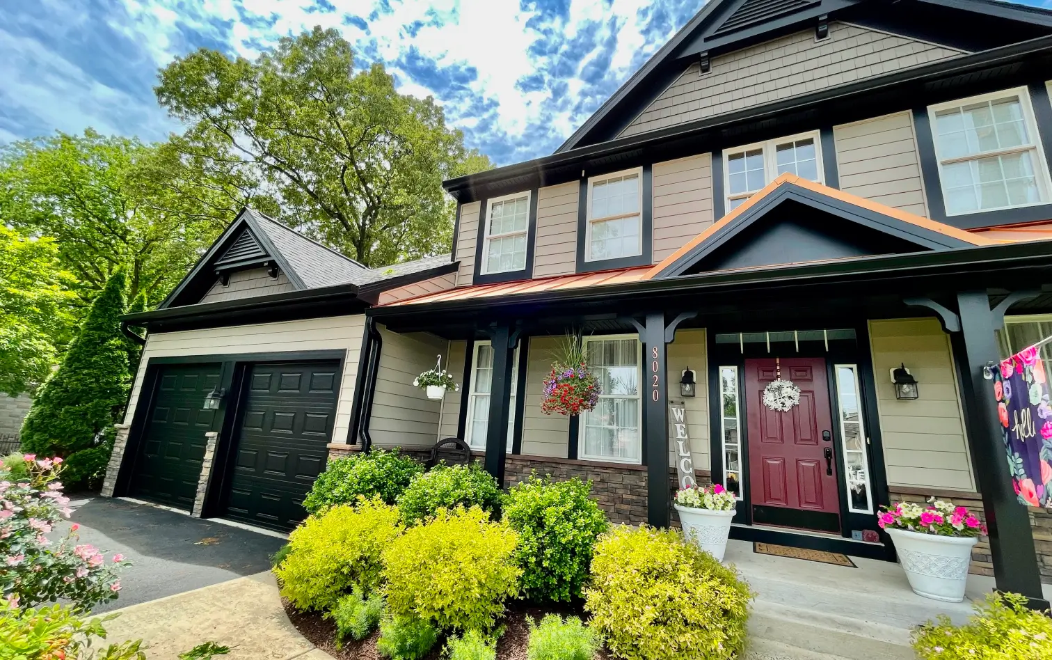 Two-story house with a front porch, red door, black garage, and hanging plants, surrounded by greenery under a cloudy sky.