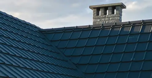 Close-up of a roof with dark shingles and a stone chimney against a cloudy sky background.