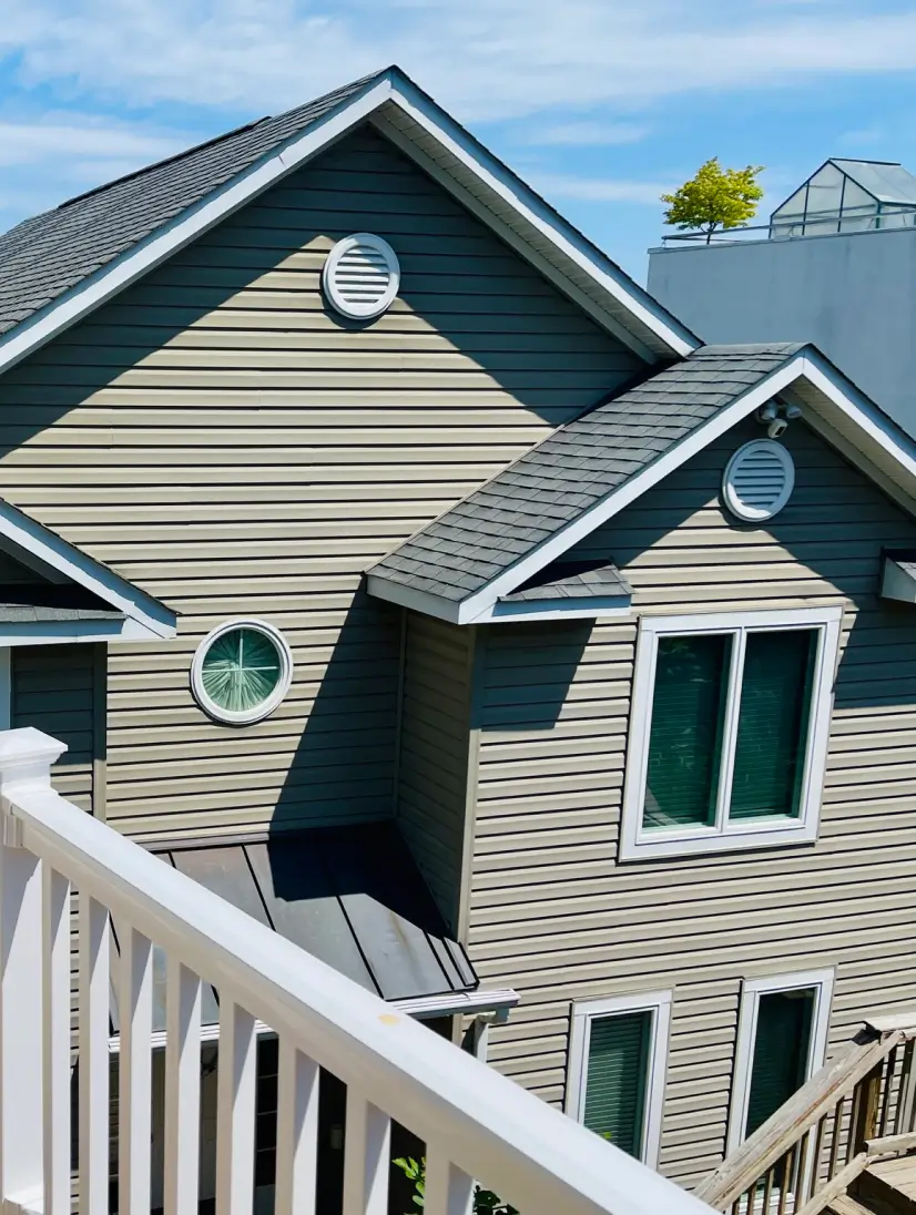 A modern house with beige siding, round windows, gray roof, and a white railing in the foreground.