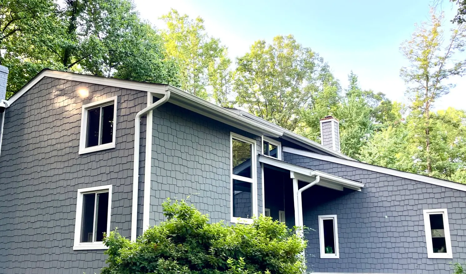 Gray house with white trim nestled among trees, featuring large windows and green shrubs in front.