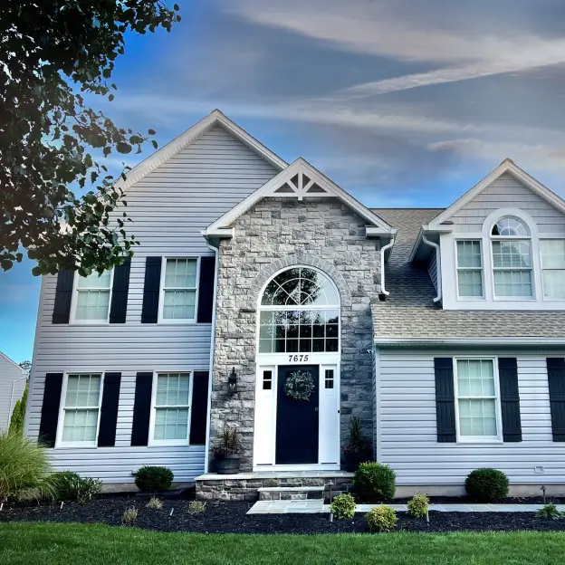 Two-story house with a stone archway, black shutters, and a small front garden under a blue sky.