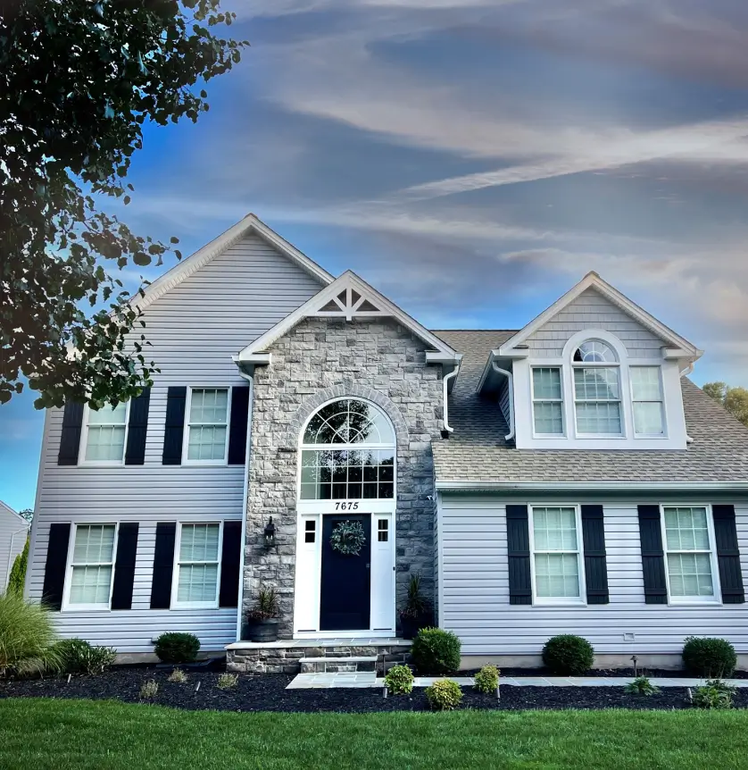 Charming suburban house with gray siding, stone facade, black shutters, and a well-maintained lawn under a blue sky.