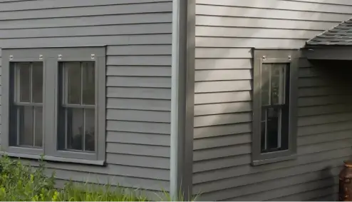 Close-up of a gray wooden house exterior with two windows and green bushes below.