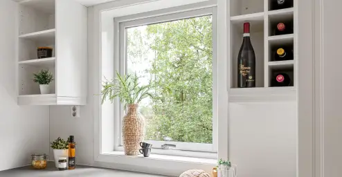 A kitchen window with a view of trees, surrounded by wine bottles, plants, and small decor items on shelves.