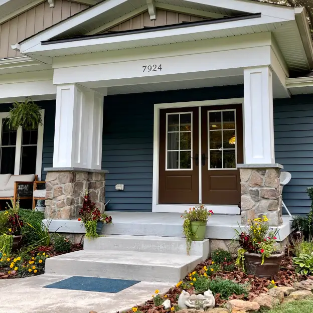 A cozy house with blue siding, a stone porch, and double doors, surrounded by potted and garden flowers.