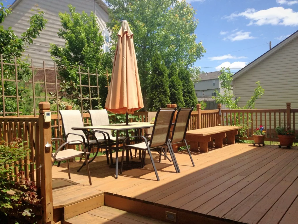 Sunny wooden deck with patio table, four chairs, a parasol, plants, and clear sky in the background.