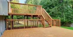 A wooden deck with stairs leading to a raised platform, surrounded by lush green trees.