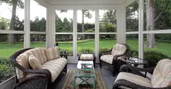 A sunroom with wicker furniture, glass coffee table, and large windows overlooking a green garden.