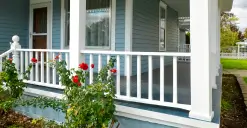 A blue house with a white porch railing and red flowers in the foreground.